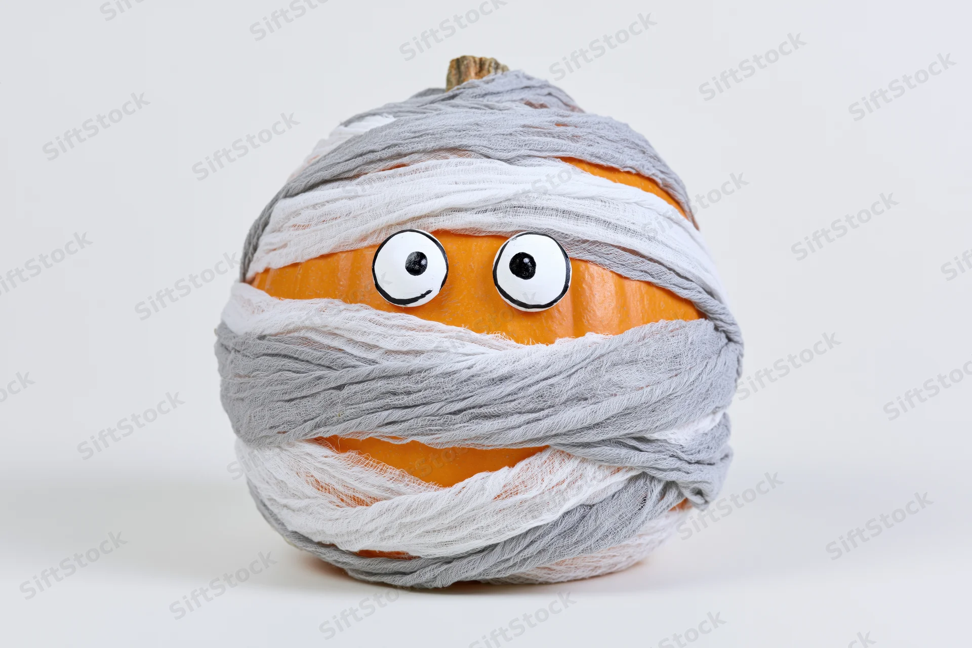 Orange pumpkin wrapped in white and gray bandages with googly eyes for halloween decoration on white background