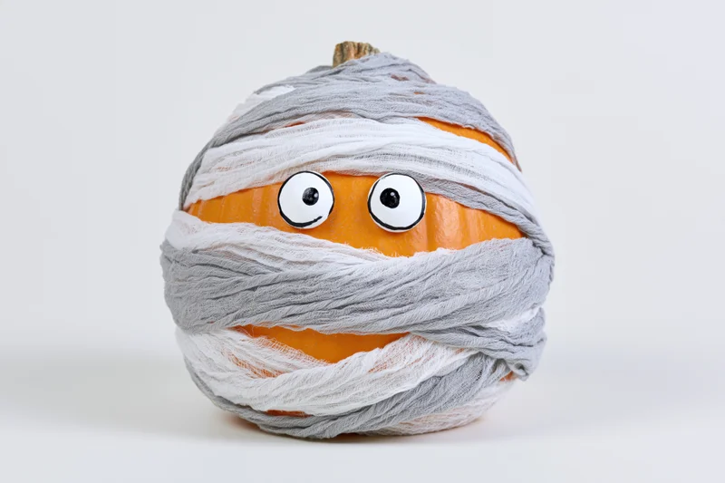 Orange pumpkin wrapped in white and gray bandages with googly eyes for halloween decoration on white background
