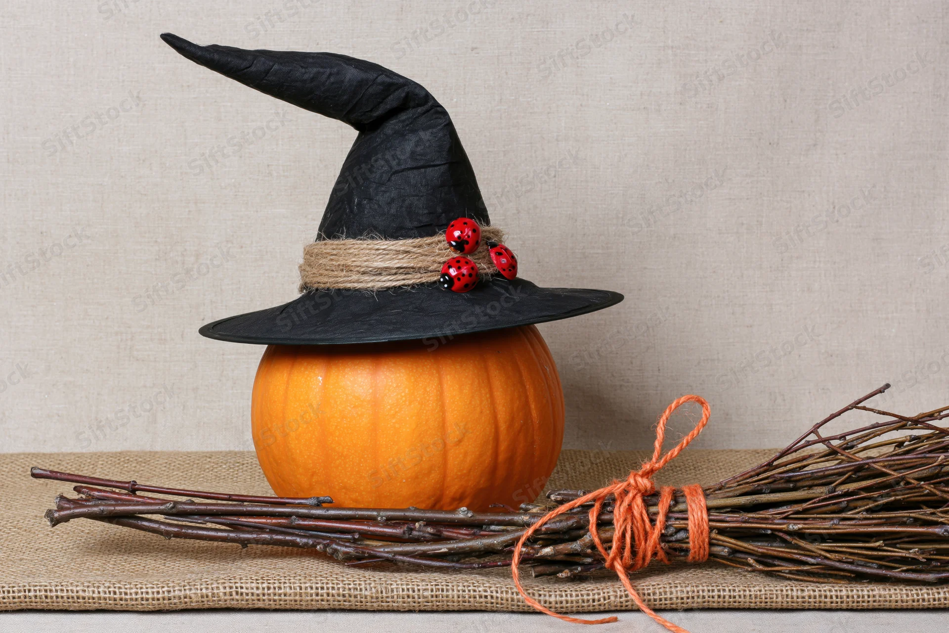 Orange pumpkin wearing black witch hat with twine and ladybugs beside a twig broom on burlap surface