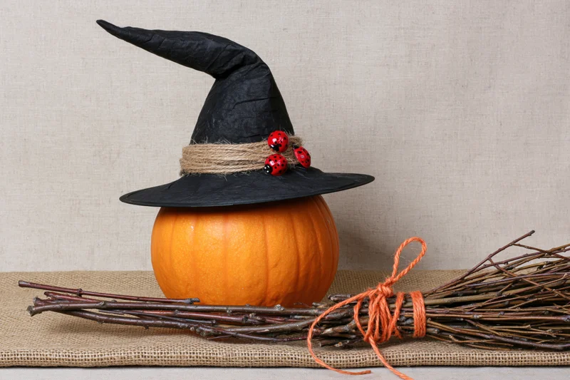 Orange pumpkin wearing black witch hat with twine and ladybugs beside a twig broom on burlap surface