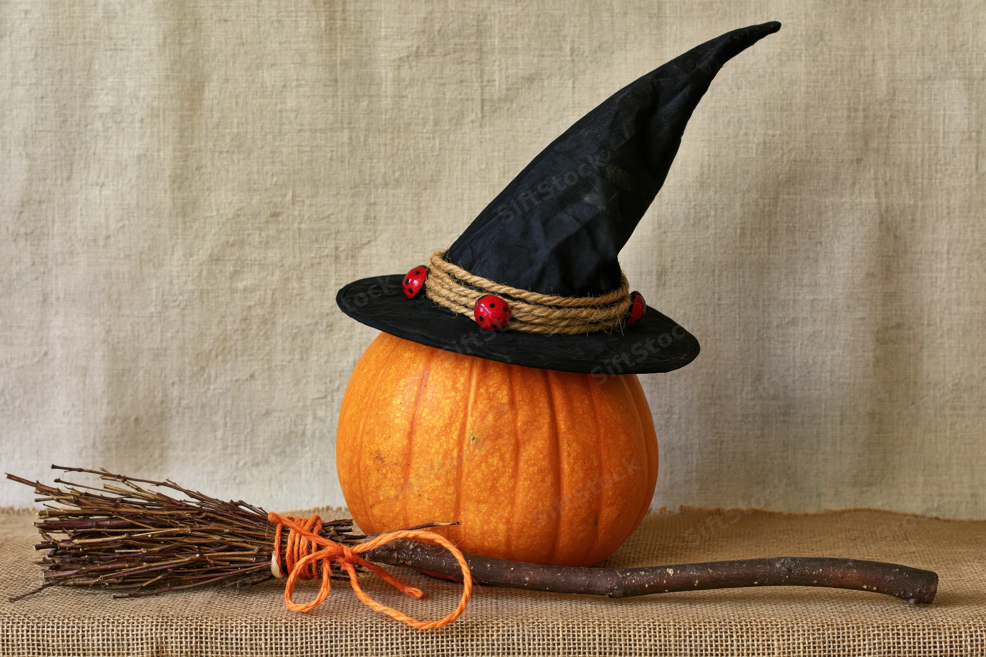 Orange pumpkin wearing black pointed witch hat with twine band and red beads, beside a twig broom on burlap fabric