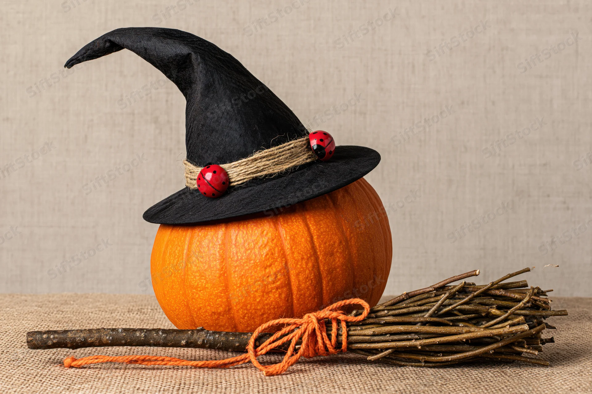 Orange pumpkin wearing black witch hat with twine band and ladybugs, beside a twig broom on burlap surface