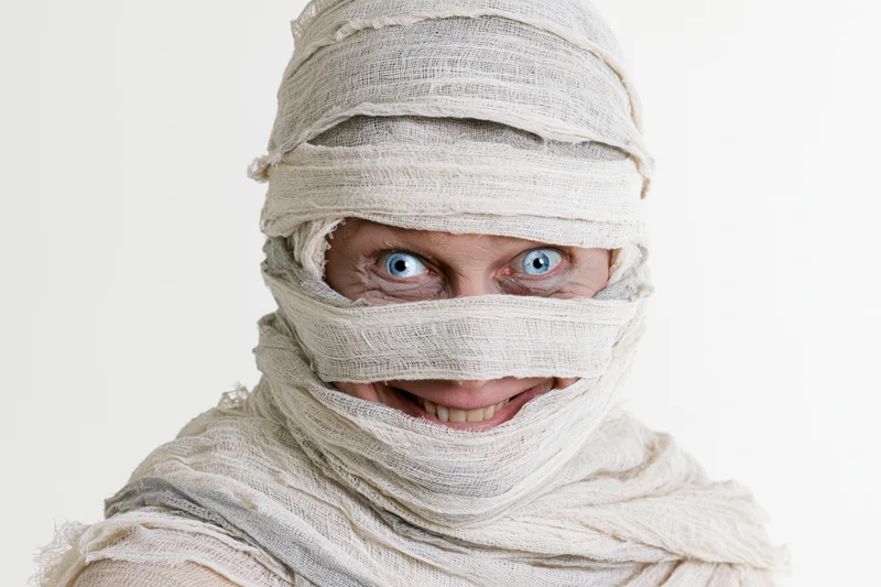 Woman dressed as a mummy with bandages wrapped around her head and face, smiling, isolated on white background