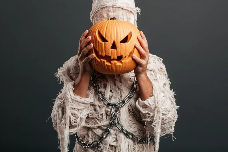 Person dressed as a mummy holding a carved jack-o-lantern in front of face against dark background