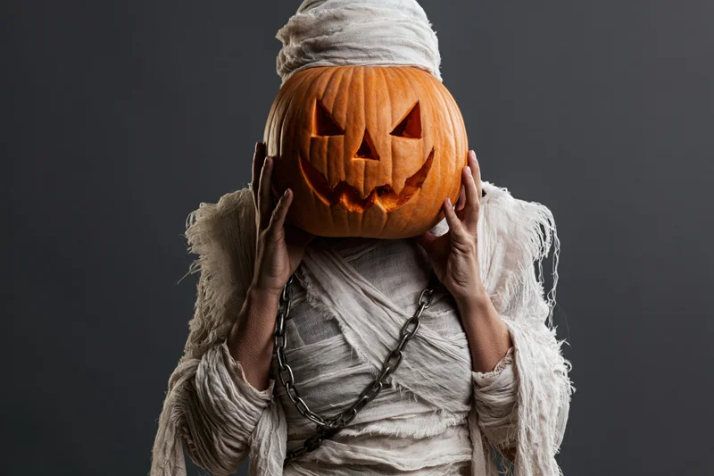 Woman dressed as a mummy holding a carved jack-o-lantern in front of her face against dark background