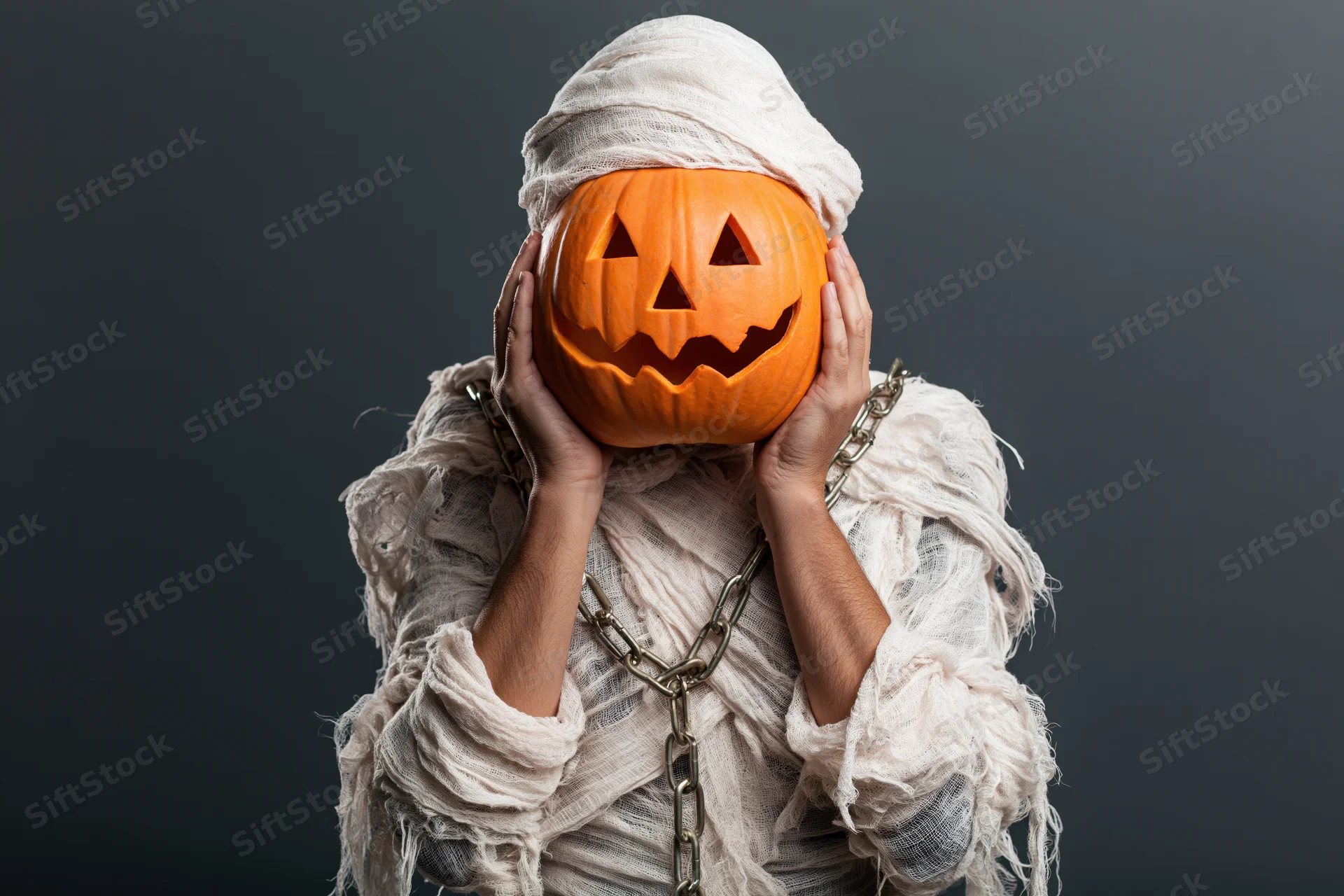 Person dressed as a mummy holding a carved jack-o-lantern in front of face against dark background