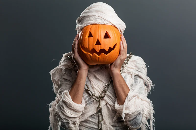 Person dressed as a mummy holding a carved jack-o-lantern in front of face against dark background