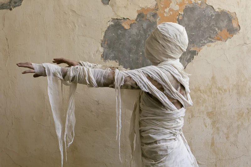 Woman in white gauze bandages reaching out against a peeling plaster wall