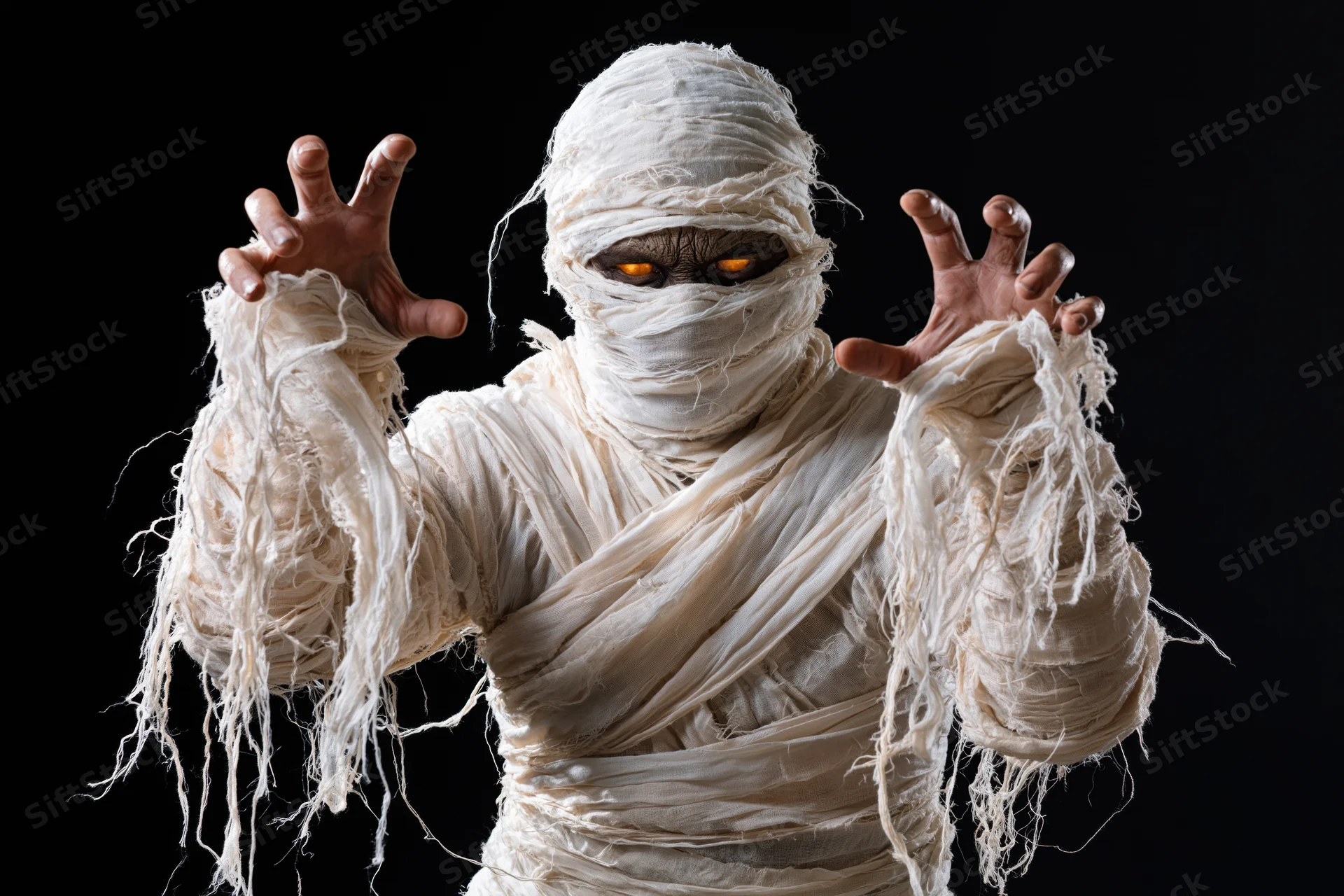 Mummy in white bandages with glowing orange eyes raising hands against black background