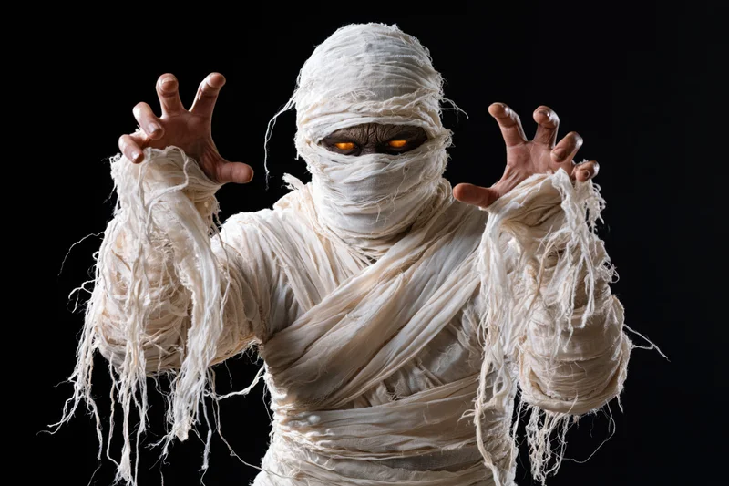 Mummy in white bandages with glowing orange eyes raising hands against black background