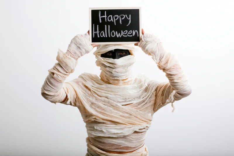 Person in mummy costume holding chalkboard sign with happy halloween message against white background
