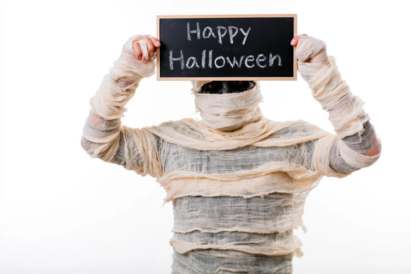 Mummy in bandages holding chalkboard sign with happy halloween text against white background
