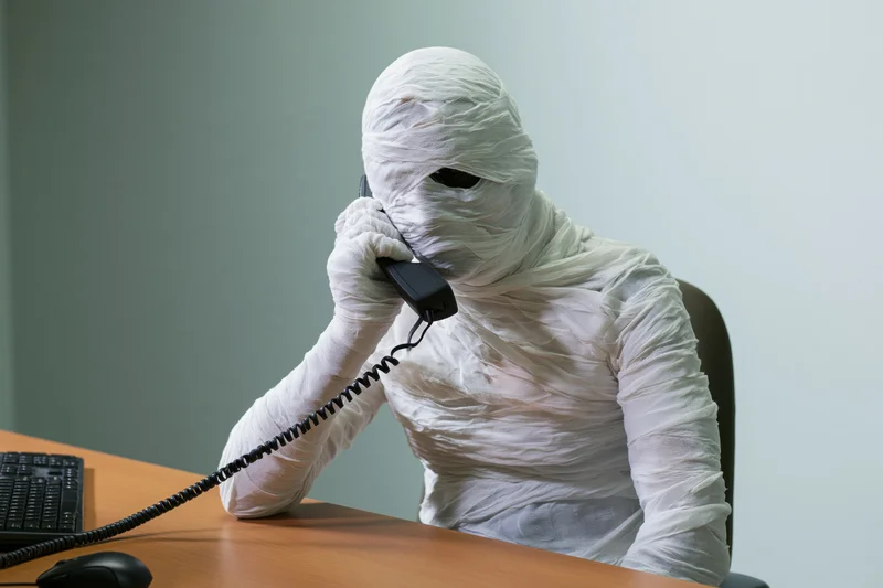 Mummy wrapped in white bandages talking on a telephone at a desk in an office