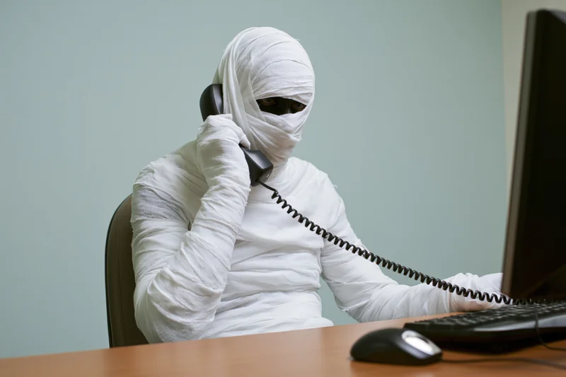 Mummy wrapped in white bandages sitting at office desk using computer and telephone