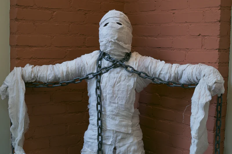 Mummy figure wrapped in white bandages with chains against a brick wall