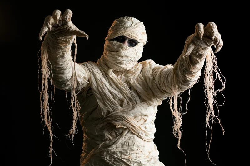 Person in mummy costume with arms raised and clawed hands against black background