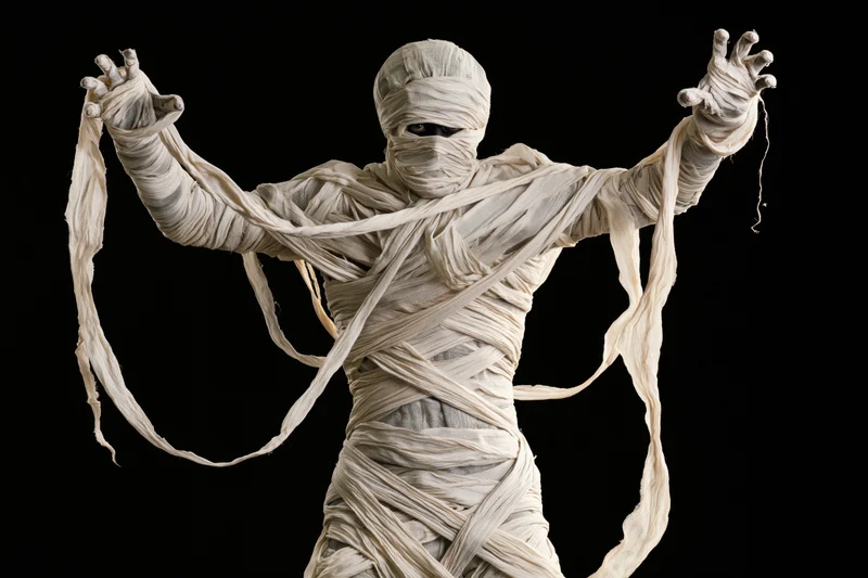 Person in full mummy costume with arms raised and hands visible against black background