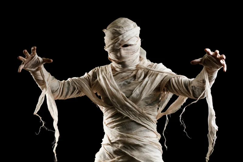 Person in full mummy costume with arms raised and hands visible against black background
