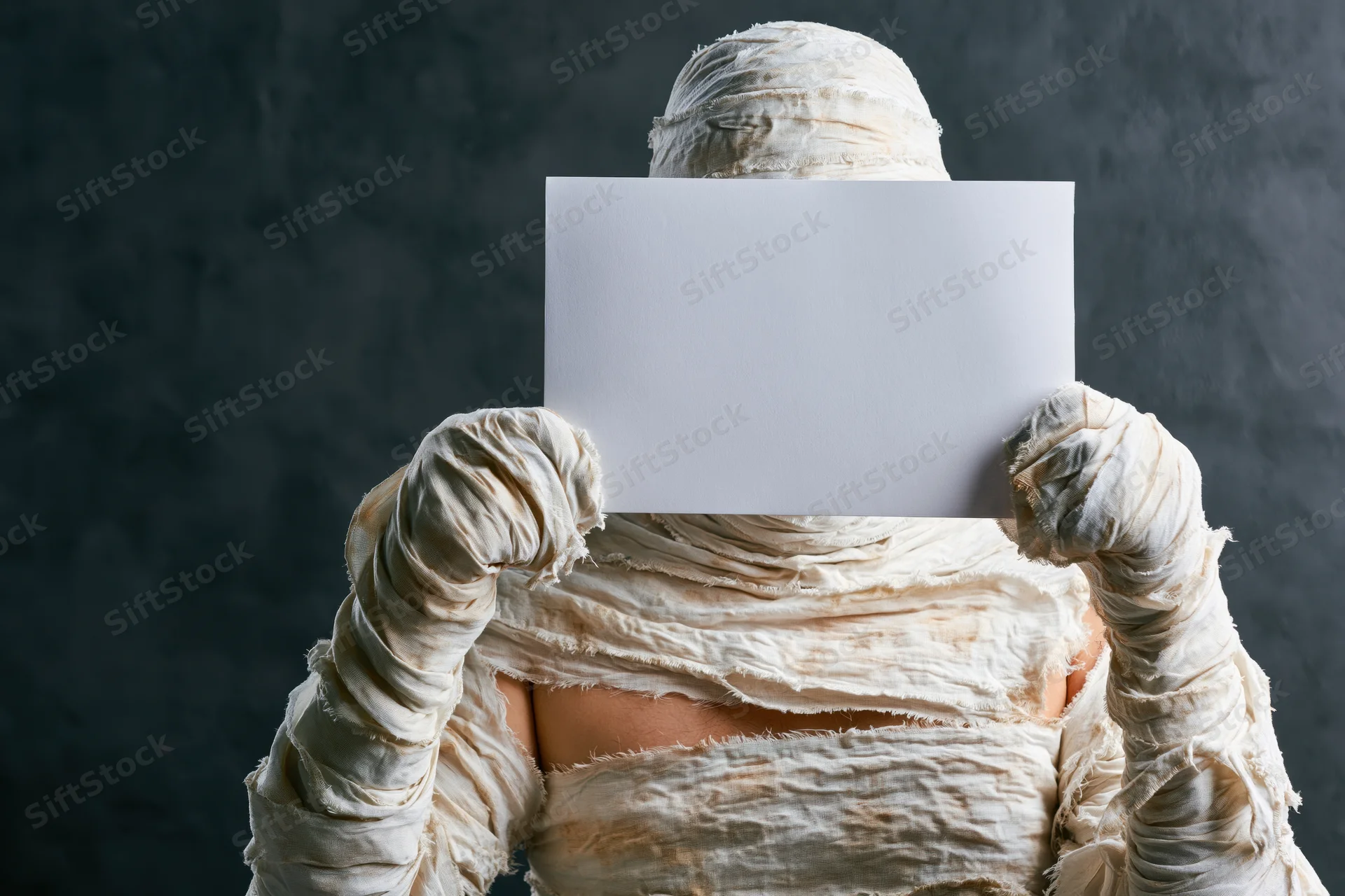 Mummy in full costume holding blank white paper in front of face against dark background