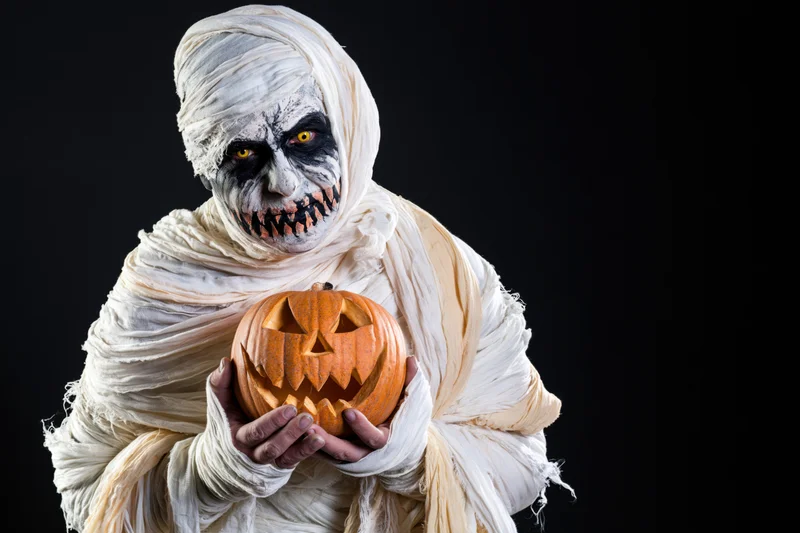 Mummy in full costume holding a carved jack-o-lantern against black background