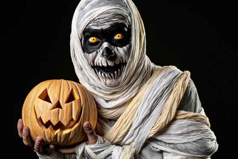 Mummy in full costume holding carved jack-o-lantern against black background