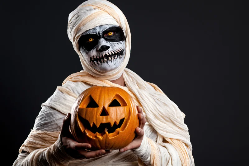 Mummy in full costume holding carved jack-o-lantern against dark background