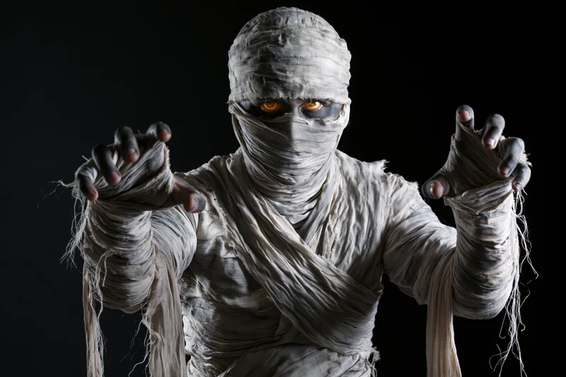 Mummy in full bandage costume with glowing eyes reaching forward against black background