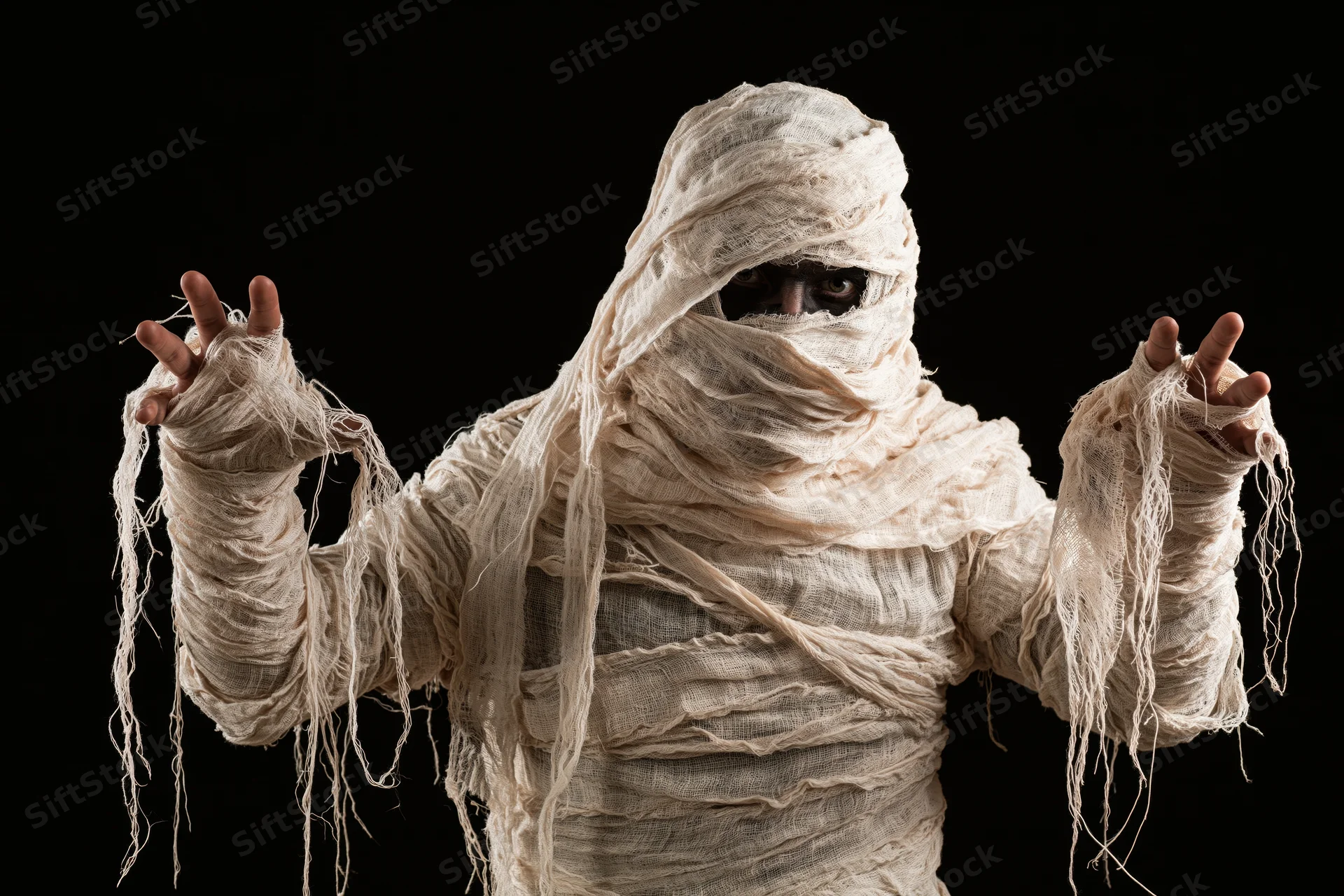 Person wrapped in white bandages as a mummy costume posing with arms raised against black background