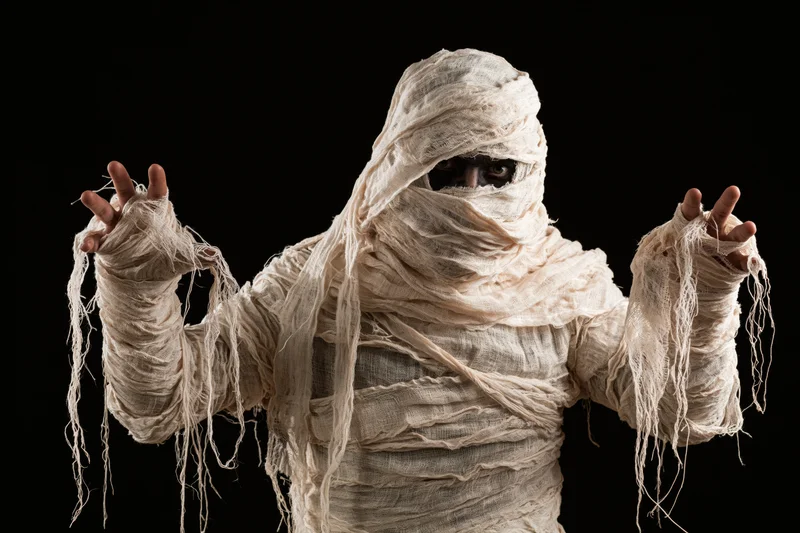Person wrapped in white bandages as a mummy costume posing with arms raised against black background