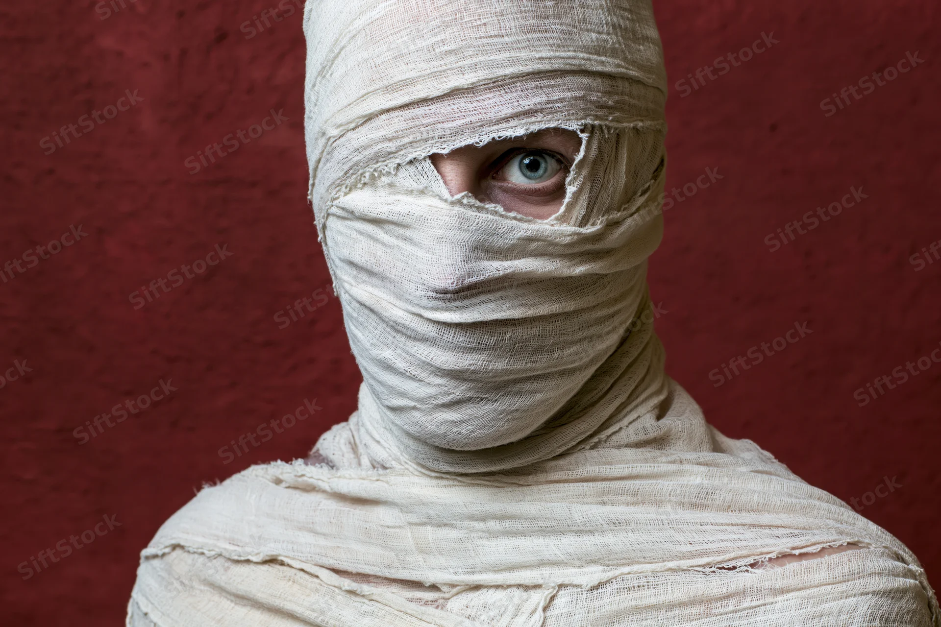 Person wrapped in white gauze bandages with one eye visible against red background