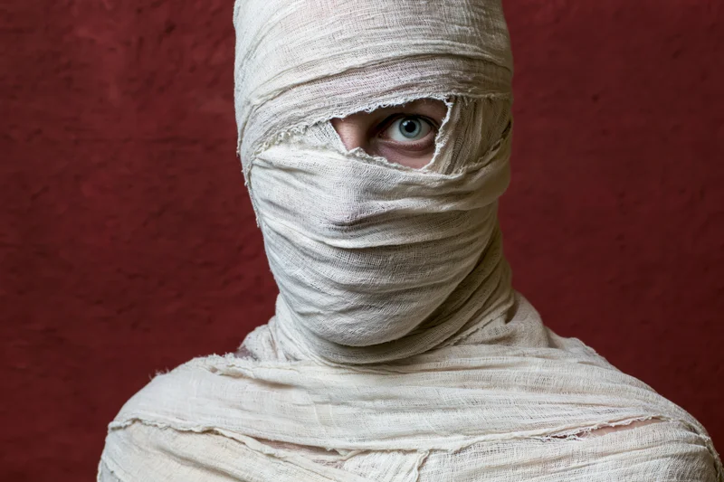 Person wrapped in white gauze bandages with one eye visible against red background