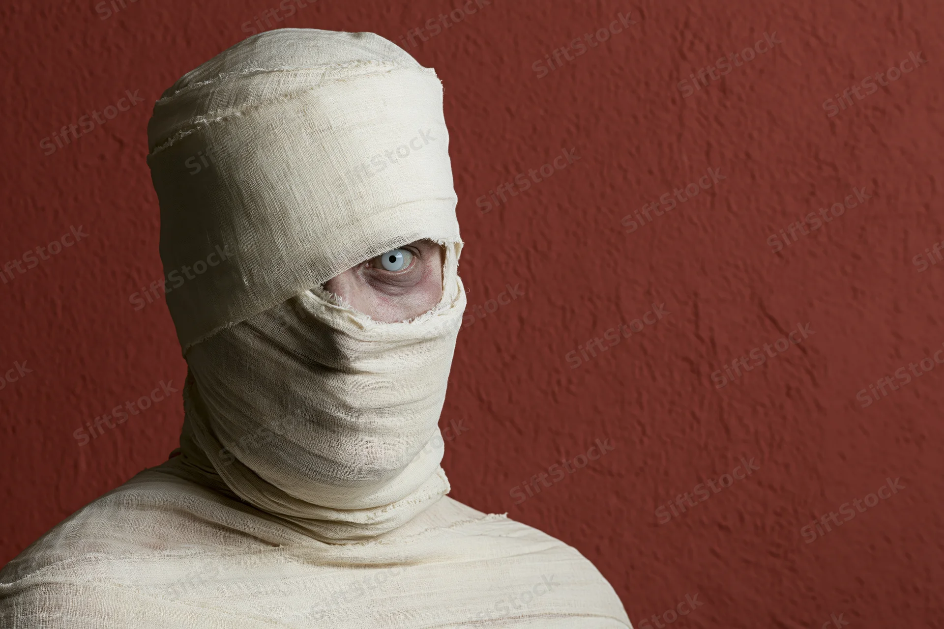 Person wrapped in gauze with one eye visible against red textured wall