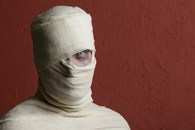 Person wrapped in gauze with one eye visible against red textured wall