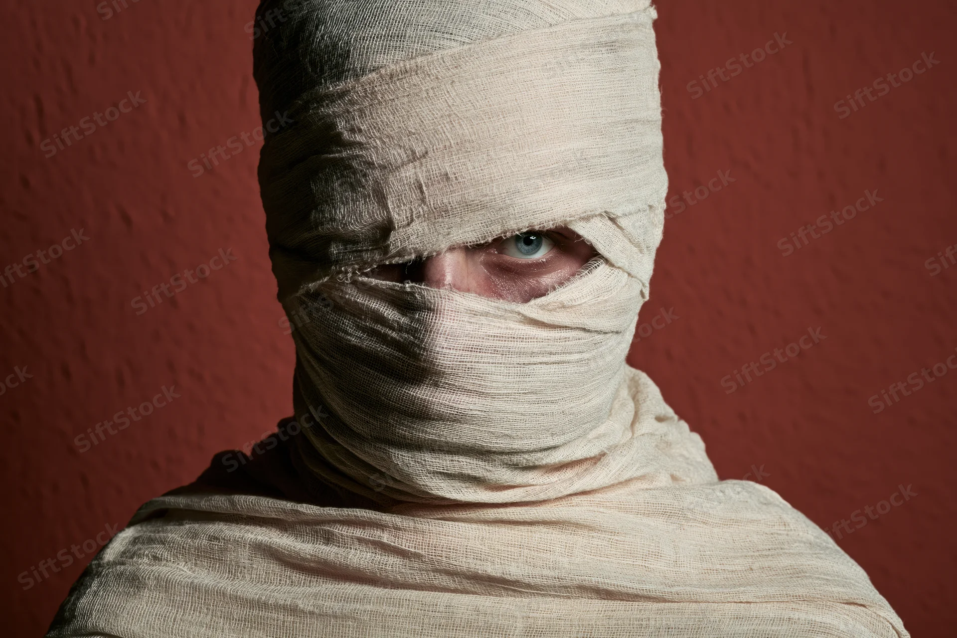 Person wrapped in medical gauze with one eye visible against red textured wall