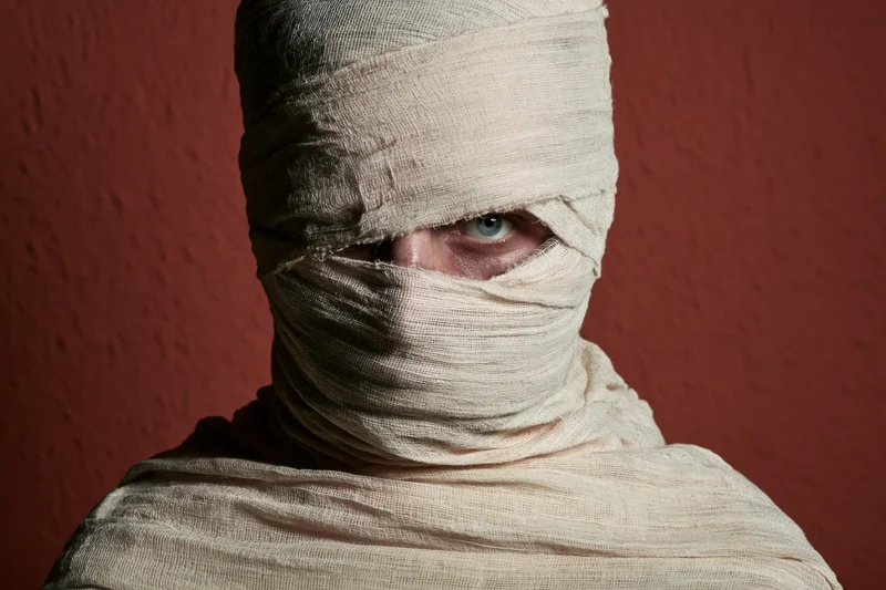 Person wrapped in medical gauze with one eye visible against red textured wall