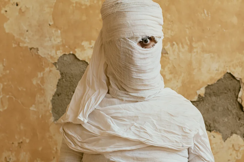 Person wrapped in white gauze with one eye visible against a peeling beige wall