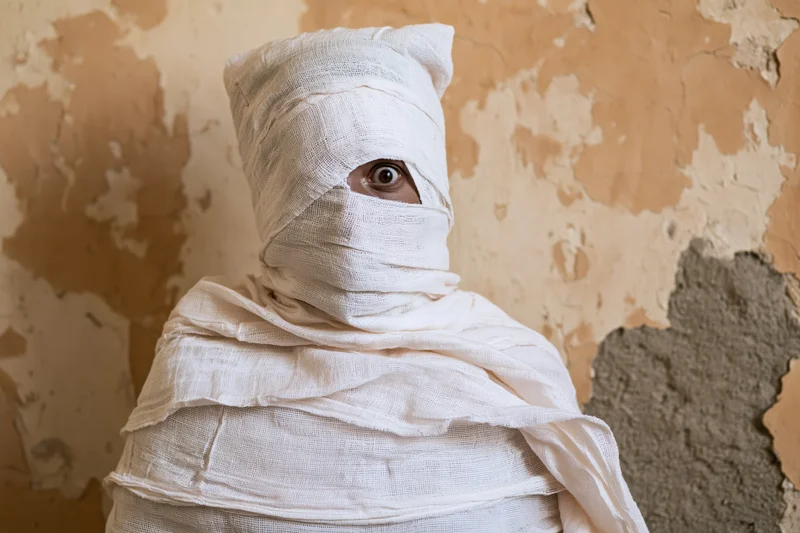Person wrapped in white gauze with one eye visible against peeling wall