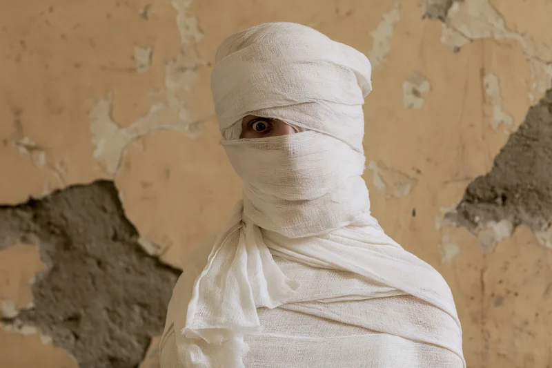 Person wrapped in white gauze bandages from head to torso against peeling wall