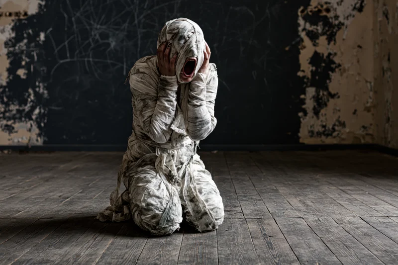Mummy wrapped in dirty white bandages screaming while kneeling on wooden floor in dark ruined room