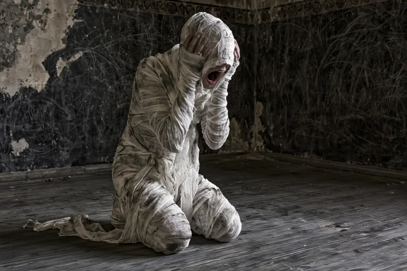 Mummy wrapped in dirty white bandages screaming while kneeling on a wooden floor in a dark crumbling room