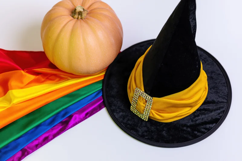 Witch hat with yellow ribbon and rhinestone buckle beside a pumpkin and rainbow pride flag on white surface