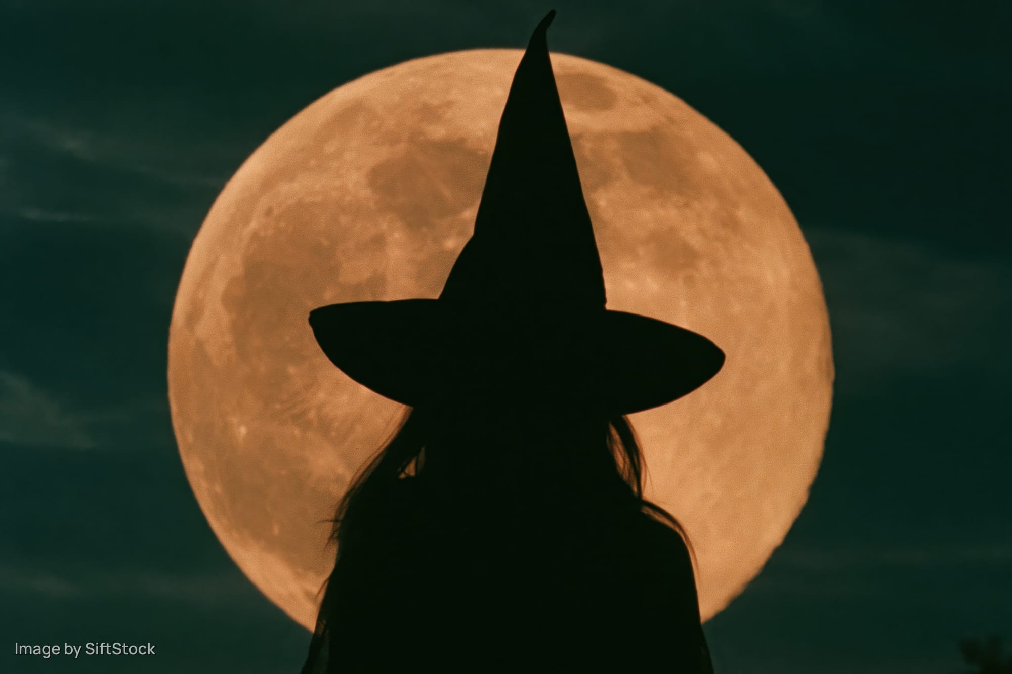 Silhouette of a woman in a pointed witch hat facing away against a full orange moon at night