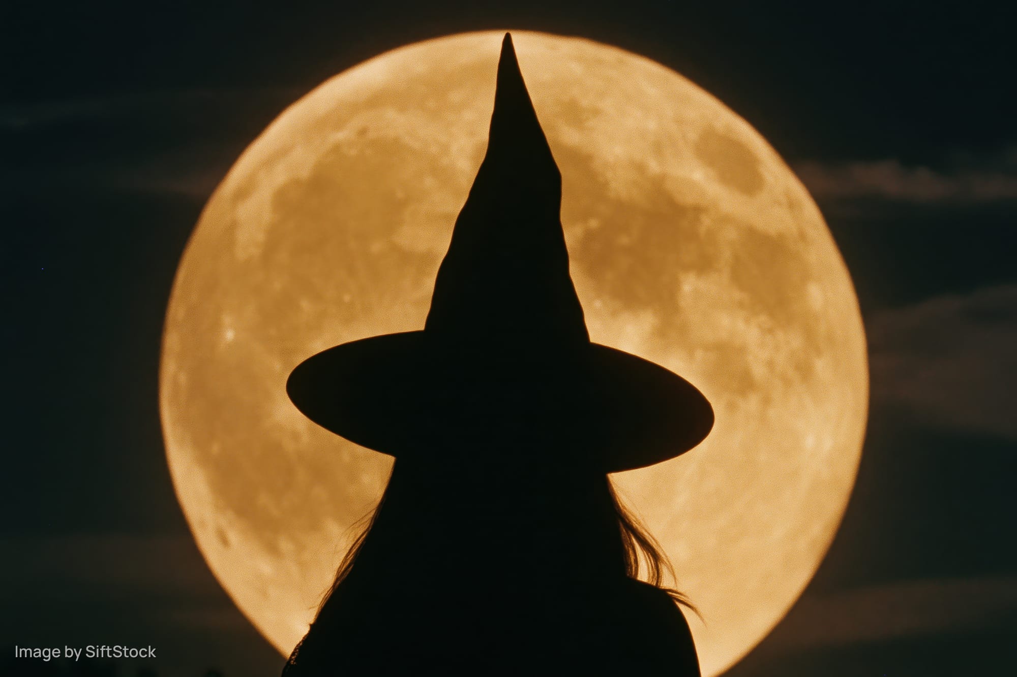 Silhouette of a woman in a pointed witch hat facing away against a full moon at night
