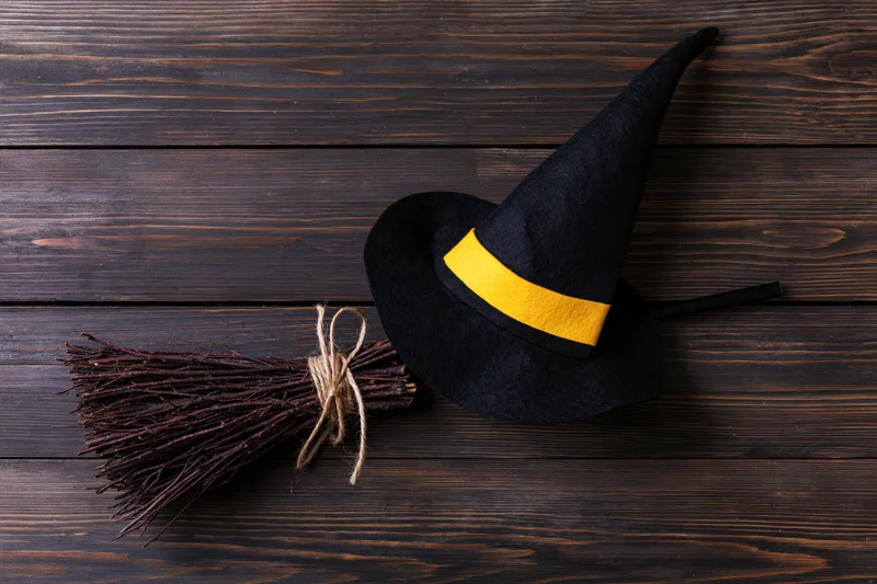 Black witch hat with yellow band and small broom on dark wooden surface