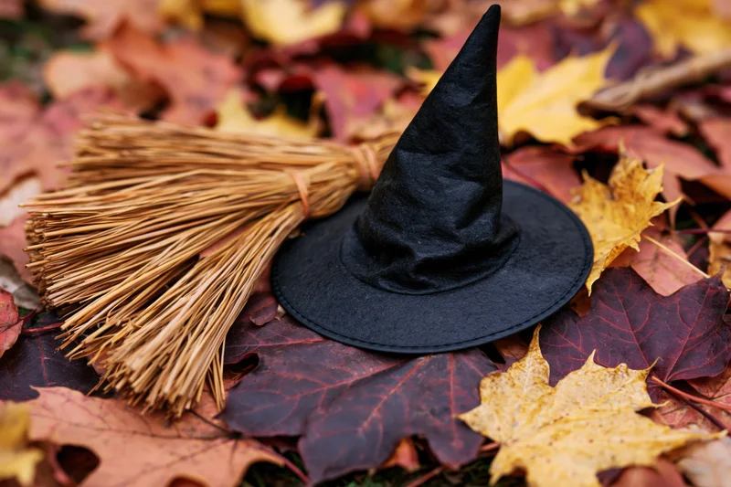 Black witch hat and miniature straw broom resting on autumn leaves