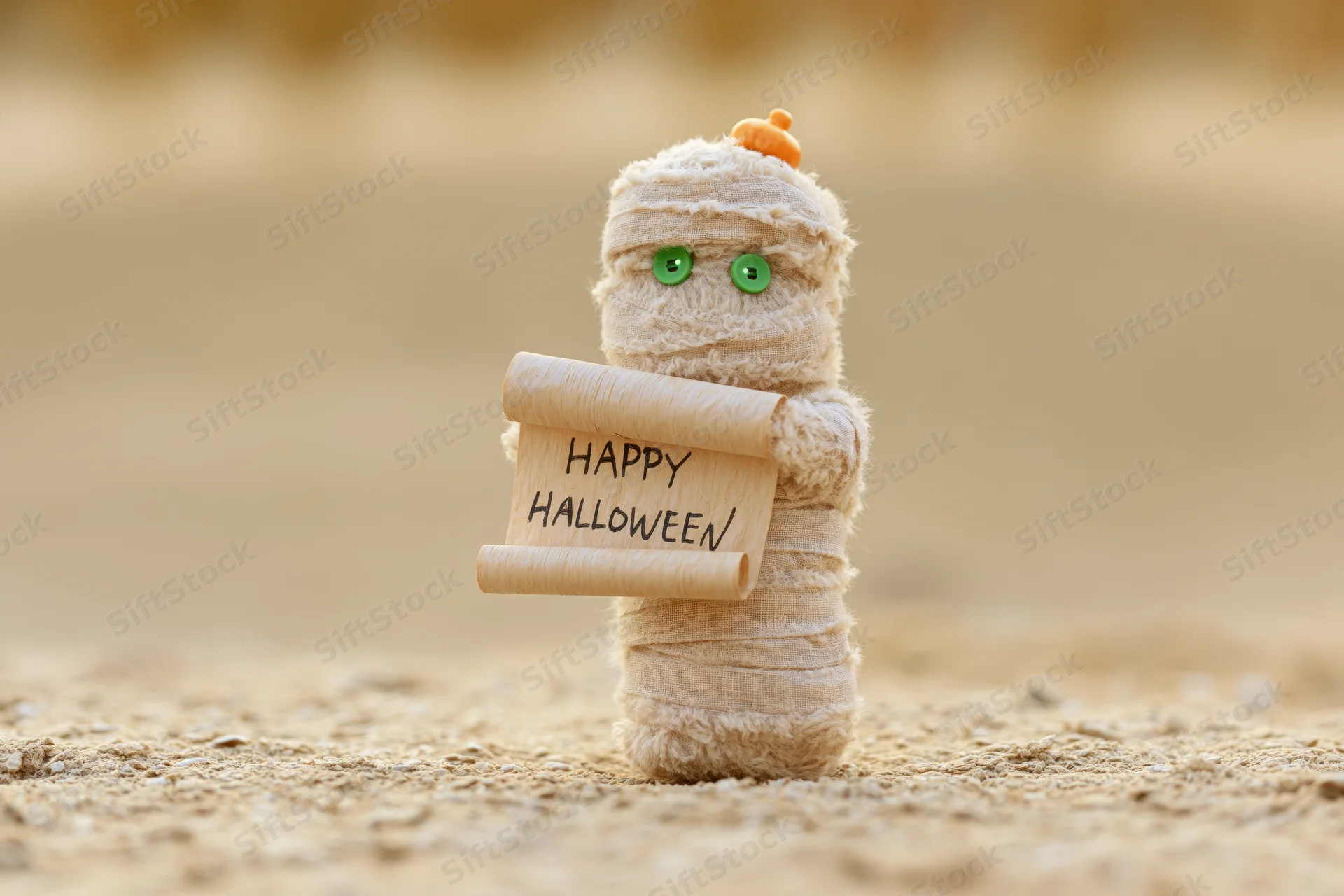Small fuzzy mummy toy holding happy halloween scroll on sandy ground