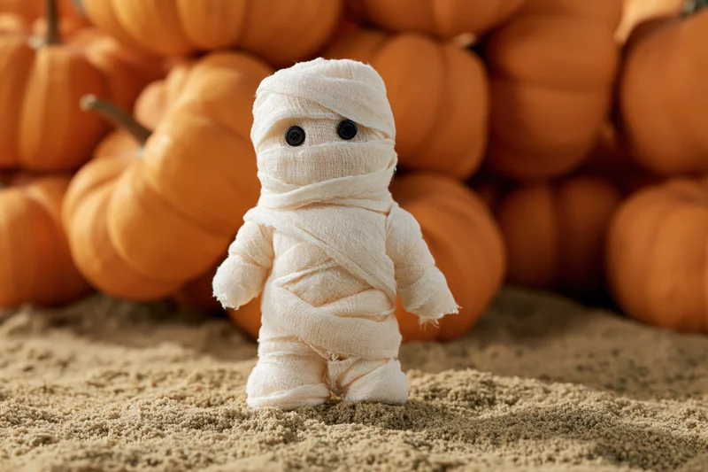 Small handmade mummy figurine wrapped in white gauze bandages standing on sand with pumpkins in background