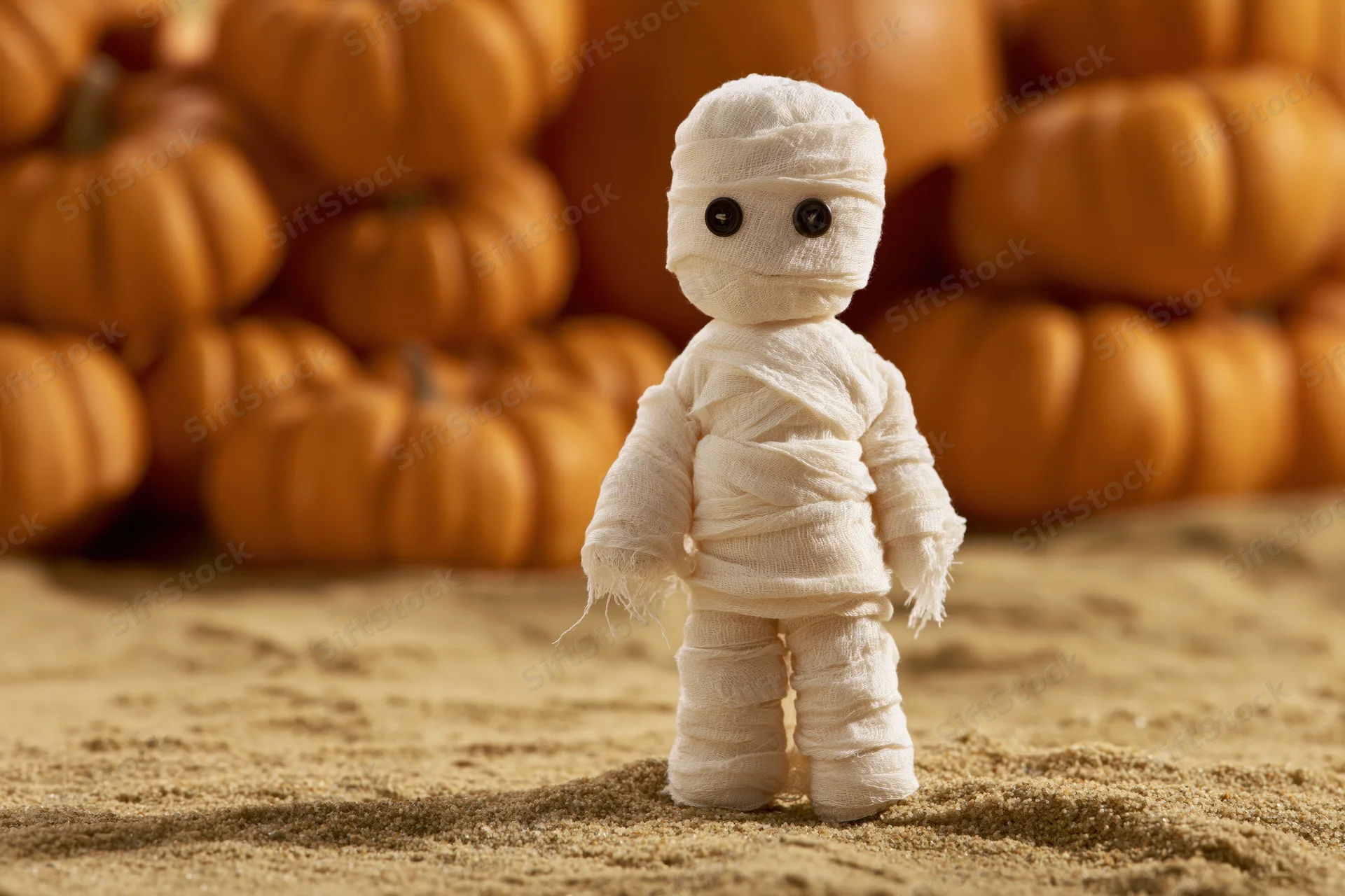 Small handmade mummy figurine wrapped in white gauze bandages standing on sand with pumpkins in background