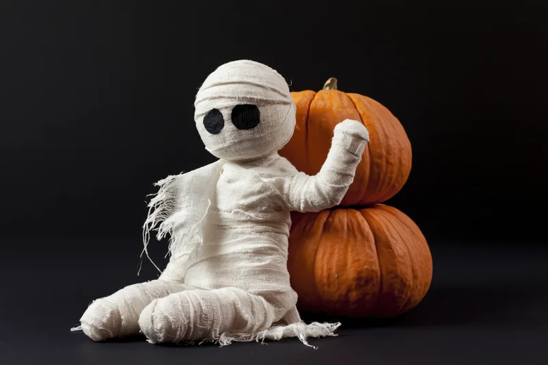 Handmade mummy figurine wrapped in white gauze bandages sitting beside stacked pumpkins on dark surface
