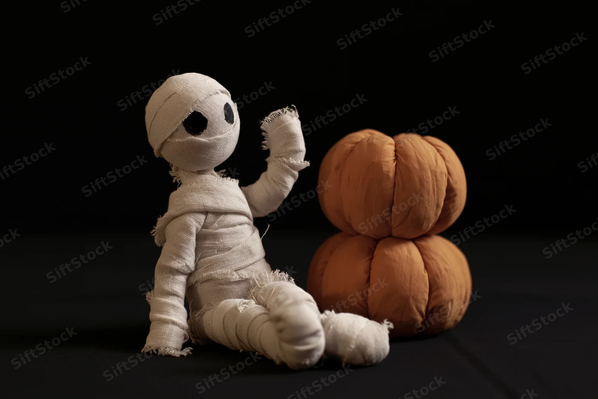Handmade mummy figurine wrapped in white gauze bandages beside a plush orange pumpkin on black background